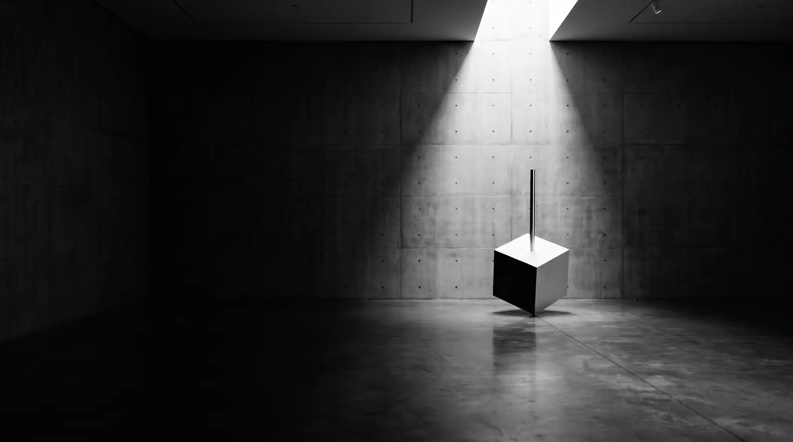 Galeria de concreto no escuro, um feixe de luz cai sobre um cubo branco atravessado por um eixo de aço